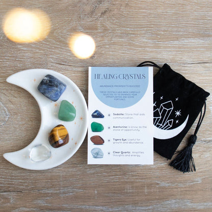 Four tumble stone crystals on a white moon-shaped trinket dish, next to a crystal information card, on a wooden table.