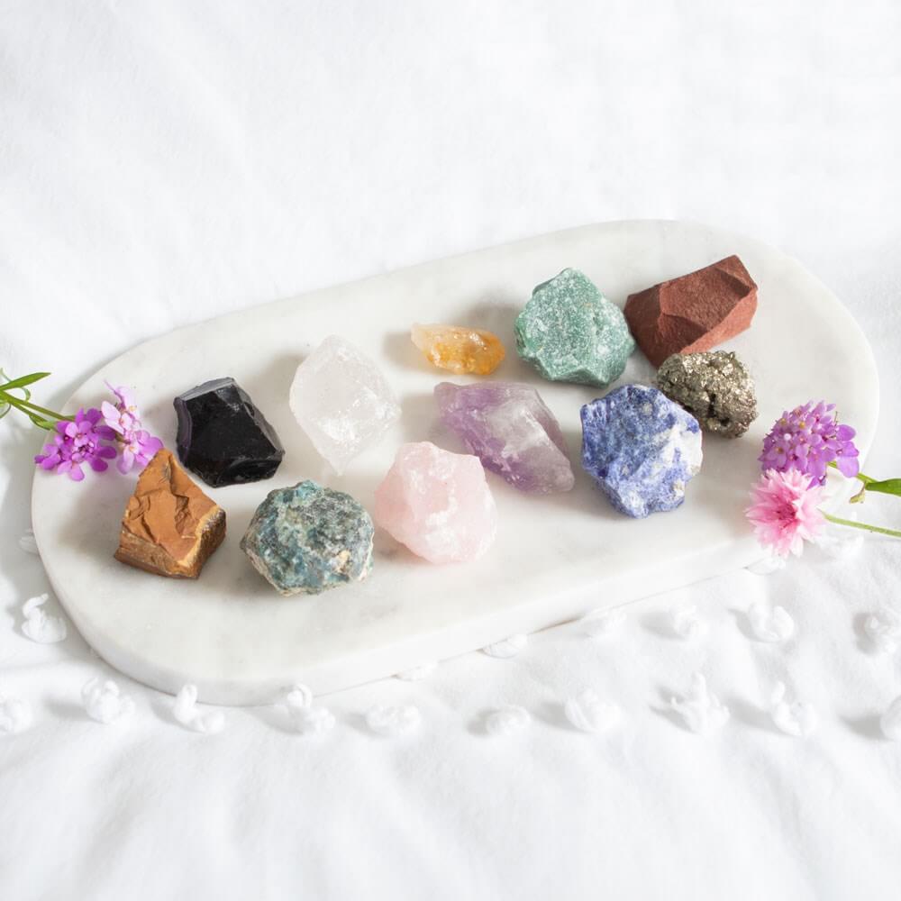 A selection of rough crystals on a large Selenite crystal dish. Best to the dish are pink and purple flowers.