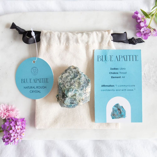 A chunk of Blue Apatite crystal on top of a cotton drawstring bag, next to an information card and two purple flowers.