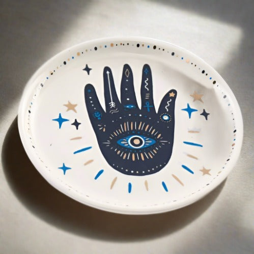 A white trinket dish that features a blue and gold Hamsa Hand Evil Eye design in the middle and magical symbols.