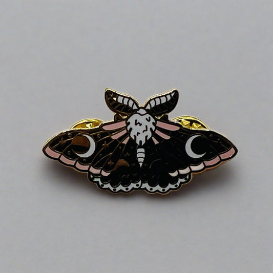 A moth pin badge brooch with blue and pink enamelled wings. In each wing there is a white crescent moon.