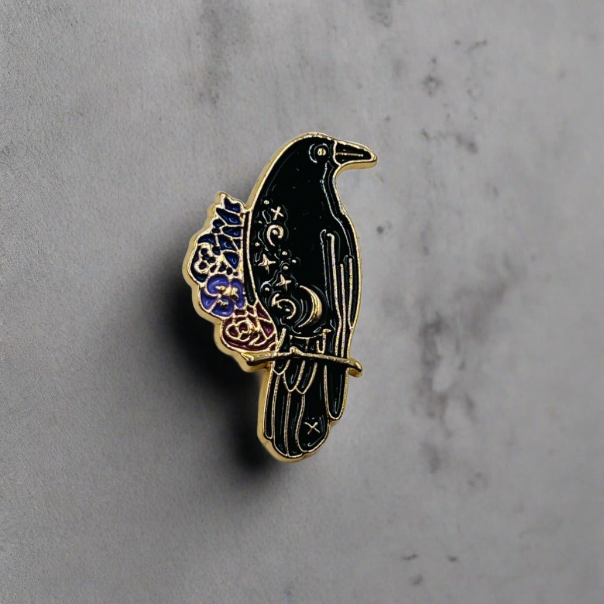 A black raven enamelled pin badge brooch. The raven is perched on a small branch and has purple flowers next to it.